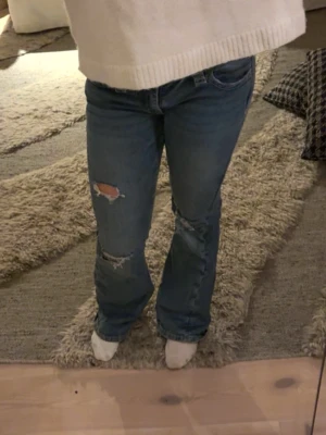 Blå bootcut jeans med slitningar - Säljer ett par blå bootcut jeans med slitna detaljer och hål på benen. Jeansen har låg midja och en snygg broderad bakficka som sticker ut. Tillverkade i klassiskt denimtyg och perfekta för en avslappnad look. Köptes på Gina Young. Dem är i bra skick men lite lite slitna nedtill. 