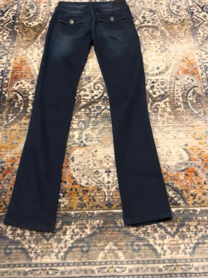 Mörkblå True Religion jeans - Säljer ett par mörkblå jeans från True Religion, modell Cora Mid Rise Straight. Jeansen har raka ben, klassiska fem fickor med snygga detaljer och silverfärgade knappar. Materialet är jeans med en skön stretch och tvättade partier för en cool look. Bara provat inte använda.