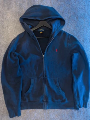 Ralph Lauren Zip Hoodie - Tröjan passar som Storlek S i herr. Mörkblå zip-hoodie från Polo Ralph Lauren med huva, dragkedja och fickor framtill. Klassisk röd Polo-logga på bröstet. Bekväm och stilren tröja i väldigt gott skick, perfekt för dig som gillar en clean, tidlös och sportig stil.