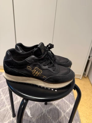 Svarta sneakers  - Snygga svarta sneakers från Gant med guldfärgad logga på sidan och svart snörning. Ovandelen är i en mix av skinn och syntet, och sulan är vit med svarta detaljer undertill. Perfekta för dig som gillar klassisk men sportig stil.