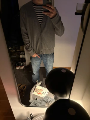 Grå kofta från Acne Studios - Säljer en stilren grå kofta från Acne Studios med knäppning framtill och diskret logopatch på bröstet. Koftan har en normal passform och är tillverkad i mjuk bomull, perfekt för lager-på-lager under kyliga dagar.