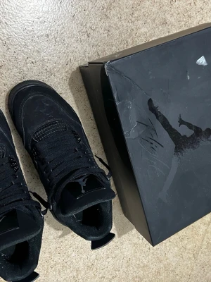 Svarta Nike Air Jordan 4 Retro - Säljer ett par helsvarta Nike Air Jordan 4 Retro med klassisk Jumpman-logga på häl och plös. Skorna har snörning, platt sula och är tillverkade i mocka och syntet. Grymma detaljer och ikonisk siluett, perfekt för dig som gillar streetwear och sneakerskultur. Kan rengöras. 