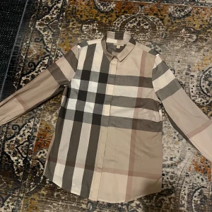 Rutig skjorta från Burberry Brit M - Säljer en klassisk rutig skjorta från Burberry Brit i storlek M. Skjortan har stora beige, bruna, svarta och vita rutor, lång ärm och knappar framtill. Tillverkad i mjuk bomull och har en snygg krage. Perfekt för dig som gillar stilrena och tidlösa plagg.