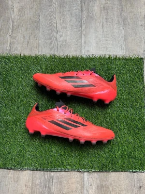Röda Adidas F50 fotbollsskor  - Säljer ett par röda Adidas F50 fotbollsskor med svarta detaljer och klassiska tre ränder på sidan. Skorna har snörning, platt sula med fasta dobbar och är tillverkade i syntetmaterial för lätt känsla på planen. Perfekta för dig som vill sticka ut på fotbollsplanen.