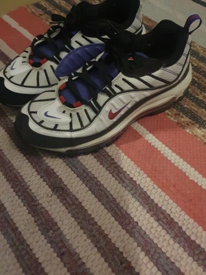 Nike Air Max 98 sneakers vit/lila/röd - Nike Air Max sneakers i vitt med svarta detaljer, lila mesh och röda accenter. Skorna har synlig Air-sula, svart snörning och rund tå. Swoosh-logga i rött och lila på sidan och tån. Materialmix av textil och syntet för en sportig look.