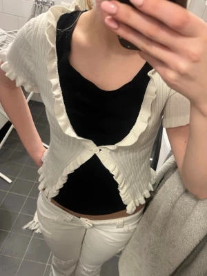 Vit ribbad kofta med volangkant - Säljer en söt vit ribbad kofta med korta ärmar och volangkant längs öppningen och nederkanten. Koftan har en knapp framtill och är croppad i modellen, perfekt att slänga över ett linne eller t-shirt.