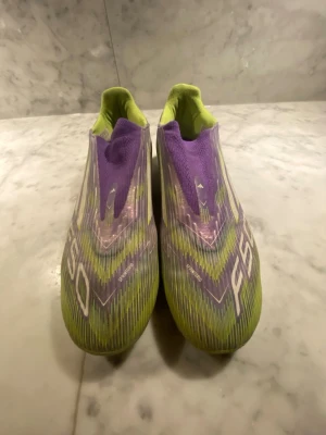 Adidas F50 lila och lime fotbollsskor - Säljer ett par Adidas F50 fotbollsskor i en riktigt snygg färgkombo av lila och limegrönt. Skorna har en strömlinjeformad design utan snörning, med Adidas-logga bak och grafiska detaljer på ovandelen. Perfekta för dig som vill sticka ut på planen. Adidas PRO 