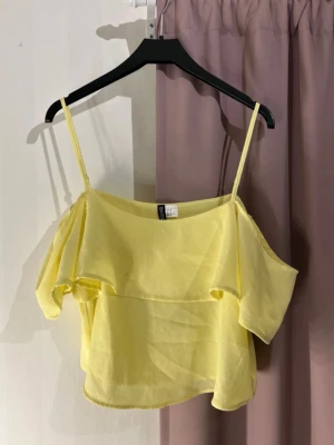 Gul offshoulder topp från H&M - Söt gul offshoulder topp från H&M med tunna axelband och volangdetaljer. Toppen är luftig och har en lätt, flowy känsla som passar perfekt till varma dagar. Tillverkad i ett tunt, mjukt material som känns skönt mot huden.