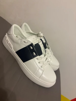 Valentino Garavani vita sneakers - Säljer ett par snygga vita Valentino Garavani sneakers i skinn med bred svart rand och coola nitar på hälen. Klassisk rund tå, vita snören och platt sula med logga undertill. Perfekta för dig som vill ha en lyxig och stilren look.