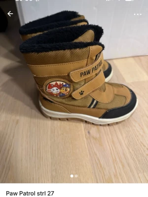 Bruna Paw Patrol vinterstövlar strl 27 - Säljer ett par bruna Paw Patrol vinterstövlar med svart foder och kardborreband. Skorna har coolt Paw Patrol-märke på sidan och slitstark sula. Perfekta för kalla dagar och lek utomhus!