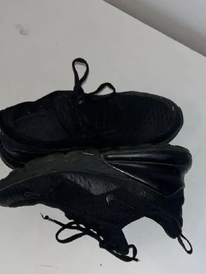 Svarta Nike Air Max 270s - Säljer ett par helsvarta Nike Air Max sneakers med klassisk luftsula och meshdetaljer. Skorna har snörning och en sportig vibe, perfekta för dig som gillar streetwear. Swoosh-loggan syns på sidan och sulan är riktigt chunky.