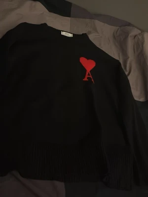 Svart stickad tröja från Ami - Säljer en svart stickad tröja från Ami Alexandre Mattiussi med ikonisk röd hjärtdetalj och bokstaven A på bröstet. Tröjan har rund halsringning och ribbade muddar. Perfekt för dig som vill ha en clean men ändå statement look.