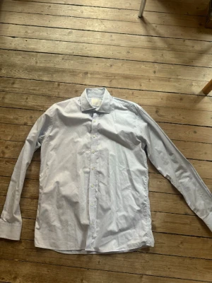 Ljusblå skjorta från Oscar Jacobson - Klassisk ljusblå skjorta från Oscar Jacobson med normal passform och långa ärmar. Skjortan har knappar framtill och är tillverkad i mjuk bomull. Perfekt till jeans eller kavaj