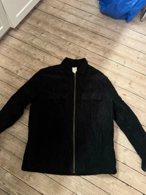 Svart manchester overshirt från H&M - Säljer en svart overshirt från H&M i mjuk manchester med dragkedja framtill och två bröstfickor. Jackan har klassisk krage och raka ärmar, perfekt för lager på lager och en clean look.