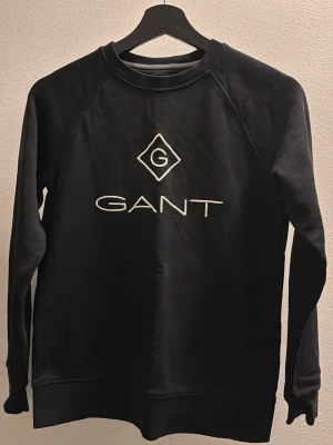 Svart sweatshirt från GANT - Svart sweatshirt från GANT med broderad logga i vitt på bröstet. Klassisk rund halsringning och långa ärmar. 