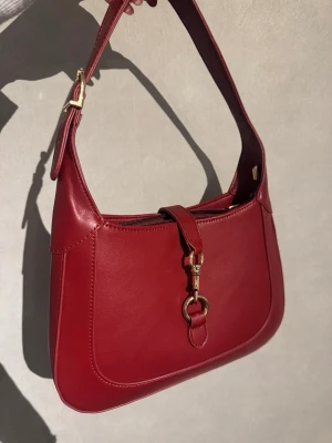 Röd axelväska från Aspry i skinn - Säljer min älskade röda axelväska från Aspry i slätt skinn med guldfärgade metalldetaljer och justerbar axelrem (Det långa axelbandet går att ta av). 