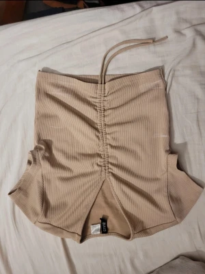 Beige ribbad croppad linnetopp - Säljer en beige croppad linnetopp från H&M med ribbad struktur och smala axelband. Toppen har snygg snörning framtill och en liten slits nedtill. Perfekt för sommardagar och enkel att styla med jeans eller kjol. Lite små vita sträck som säkert går bort i tvätt.