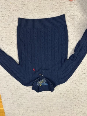 Marinblå kabelstickad tröja Polo Ralph Lauren - Kabelstickad marinblå tröja från Polo Ralph Lauren med röd broderad logga på bröstet. Tröjan har hög krage med half zip-dragkedja och ribbade muddar. Perfekt för dig som gillar klassisk och stilren look.