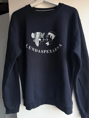 Marinblå Lundaspexarna-tröja - Mörkblå sweatshirt från Tee Jays i storlek M med tryck av Lundaspexarna på bröstet. Klassisk rund halsringning och långa ärmar. Tillverkad i mjuk bomullsmix, perfekt för chill dagar eller studenthäng.
