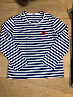 Randig långärmad topp Comme des Garçons - Säljer en ikonisk långärmad topp från Comme des Garçons PLAY i blått och vitt med klassiska ränder. Toppen har ett rött hjärta med ögon broderat på bröstet och är gjord i mjuk bomull. Perfekt för dig som gillar streetstyle och vill ha något som sticker ut.XL i barn(170) passar XS