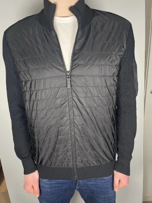 J Lindberg cardigan  - Snygg svart pufferjacka med stickade ärmar och hög krage. Jackan har quiltad framsida i glansigt material och dragkedja hela vägen. Perfekt för dig som vill ha en modern och stilren look.