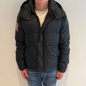 Denim & supply jacka  - Svart pufferjacka från Ralph Lauren med huva och amerikansk flagga på vänster ärm. Jackan har stora fickor med knappar och är tillverkad i polyester. Perfekt för kalla dagar och har en schysst form.