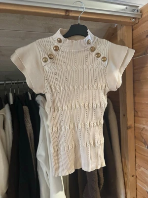 Beige stickad topp med guldknappar - Säljer en beige stickad topp med korta ärmar och ribbad hög krage. Framsidan har dekorativa guldknappar på axlarna och ett snyggt strukturerat mönster. 