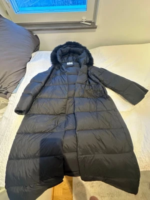 Svart lång dunjacka FUR Stockholm - Lång svart dunjacka från FUR Stockholm med luva och fuskpälskant. Jackan har bälte i midjan för snygg passform och quiltad design. Perfekt för kalla vinterdagar och riktigt mysig att bära. Klassisk look som funkar till allt. Inga hål eller skador på jackan,  Storlek S! Pris kan prutas!