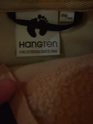 Beige fleecekofta från Hang Ten - Säljer en beige fleecekofta från Hang Ten med marinblå detaljer och dragkedja framtill. Koftan har två fickor och lång ärm, perfekt för kyliga dagar. Enkel och clean design som funkar till det mesta.