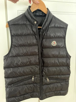 Svart dunväst från Moncler - Säljer en svart dunväst från Moncler med klassisk quiltad design och hög krage. Västen har två dragkedjeförsedda fickor framtill och ikonisk Moncler-logga på bröstet. Perfekt att slänga över en hoodie eller tröja för extra värme.