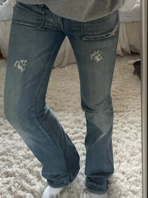 Blå bootcut jeans från Diesel - Säljer ett par skitsnygga blå bootcut jeans från Diesel med slitna detaljer på låren och coola fickor. Jeansen är lågmidjade och sitter bra på mig som är 168 cm. 