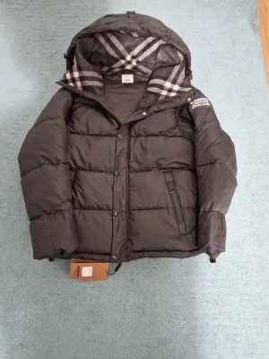 Svart pufferjacka från Burberry - Svart pufferjacka från Burberry med klassiskt rutigt foder i huvan. Jackan har dragkedja och knappar framtill, samt fickor med dragkedja. Perfekt för kyliga dagar och har en modern, boxig passform.