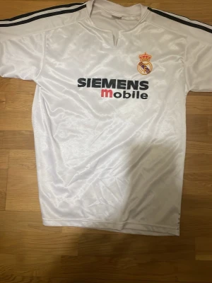 Real Madrid Zidane 5 tröja vit - Vit Real Madrid tröja med svarta och orange detaljer, Siemens Mobile-tryck på bröstet och Zidane 5 på ryggen. Klassiska svarta ränder på axlarna och klubbmärke på bröstet. Tillverkad i blankt syntetmaterial, perfekt för fotbollsfans.