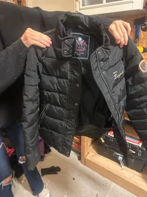 Svart pufferjacka Horse Legend - Säljer en svart pufferjacka från Horse Legend Wear med broderad logga på bröstet och ärmen. Jackan har hög krage, dragkedja och tryckknappar framtill samt två fickor med dragkedja. Perfekt för dig som gillar stilren och sportig look.