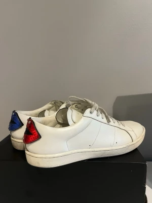 Saint Laurent vita sneakers med läppar - Unika vita sneakers från Saint Laurent med coola läppar i blått och rött på hälen. Skorna har snörning, rund tå och är gjorda i skinn. Märkeslogga syns både på plösen och på läpparna bak. Perfekta för dig som vill sticka ut med en statement-detalj. Storlek 41 men sitter lite större mer åt 41,5 hållet. Skorna är autentiskt och ny pris ligger på 7k!