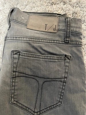 Grå jeans från Tiger of Sweden - Säljer ett par grå jeans från Tiger of Sweden med raka ben och klassisk femficksdesign. Jeansen har snygga kontrastsömmar och patch med logga bak i midjan. Materialet är mjukt jeans-tyg som sitter skönt och passar till många olika stilar.
