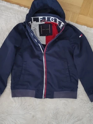 Blå vindjacka Tommy Hilfiger strl 140 - Säljer en marinblå vindjacka från Tommy Hilfiger med huva och dragkedja framtill. Jackan har snygga röda och vita detaljer längs dragkedjan och logga på ärmen. Insidan har meshfoder och tryckt Hilfiger-text. Perfekt för dig som vill ha en stilren och sportig look.