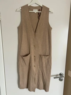 Beige lång stickad väst Holly & White - Lång beige stickad väst från Holly & White med v-ringning, stora framfickor och knappar framtill. Enkel och stilren design som passar perfekt över en skjorta eller t-shirt. Västen har en rak passform och är gjord i mjukt material.