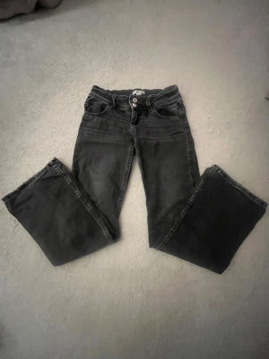 Svarta bootcut jeans med dubbla knappar - Säljer ett par svarta bootcut jeans med dubbla knappar i midjan och klassiska fem fickor. Jeansen har en lätt utsvängd passform nedtill och är tillverkade i mjukt denimtyg. Perfekta för dig som gillar en retro vibe och vill ha något som sticker ut lite extra.