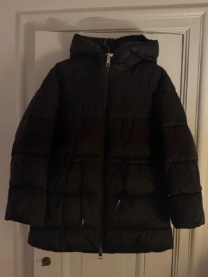 Svart pufferjacka med huva och snörning - Säljer en svart pufferjacka med huva och dragkedja framtill. Jackan har snörning i midjan för att justera passformen och är quiltad för extra värme. Perfekt för kalla dagar och har en clean, oversized look.