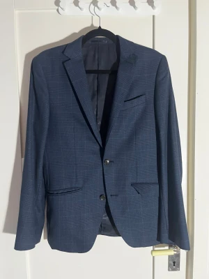 Blå rutig kavaj från Riley - Snygg blå kavaj från Riley med diskret rutmönster och två knappar framtill. Klassisk passform med fickor och slits bak. Fodrad med ljusblått tyg för extra stilpoäng. Perfekt för dig som vill ha en clean och dressad look.