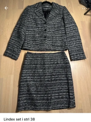 Gråmelerat kavajset från Lindex - Säljer ett snyggt tvådelat set från Lindex i storlek 38. Setet består av en gråmelerad kavaj med knappar och matchande kjol. Materialet är tjockt och strukturerat, perfekt för dig som gillar klassisk stil med modern twist.
