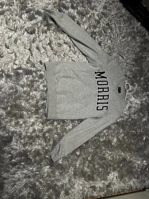 Grå sweatshirt från Morris - Säljer en grå sweatshirt från Morris med stor svartvit logga över bröstet. Tröjan har rund halsringning, ribbade muddar och är tillverkad i mjuk bomull. Perfekt för en chill och stilren look.