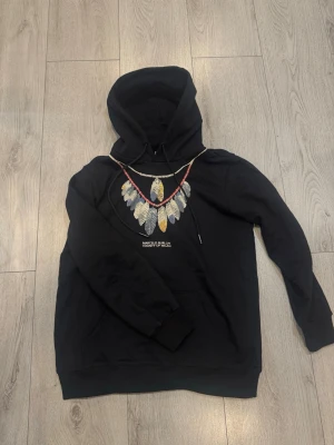 Marcelo Burlon Hoodie - Svart hoodie från Marcelo Burlon County of Milan med ikoniskt fjädertryck framtill i grå, vita och guldfärgade toner med röda detaljer samt logotyptext på bröstet. Klassisk modell med huva och dragsko, känguruficka, ribbade muddar och nederkant. Enfärgad baksida med liten röd korsdetalj på huvan, tillverkad i mjuk och bekväm bomullsblandning. Storlek M men lite större passform