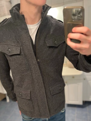 Mörkgrå ullrock från F&F Signature - Säljer en stilren mörkgrå ullrock från F&F Signature i storlek S. Rocken har fyra fickor med lock och knappar, axelklaffar och hög krage. Tillverkad i ren ull för extra värme och klassisk look. Perfekt för dig som vill ha en tidlös och snygg jacka.