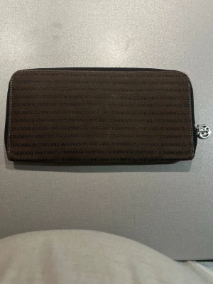 Brun plånbok från Emporio Armani - Snygg brun plånbok från Emporio Armani med logotypmönster över hela ytan och silverfärgad dragkedja. Insidan har flera fack för kort och en dragkedjeficka för mynt. Tillverkad i Italien och i ett tåligt material som känns lyxigt.
