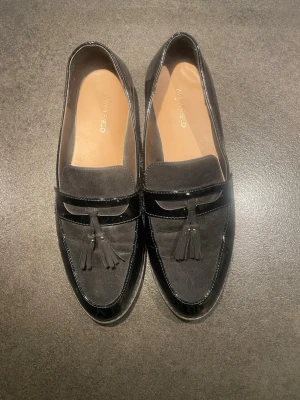 Svarta loafers från Anna Field - Snygga svarta loafers från Anna Field med dekorativa tofsar framtill. Skorna har en mix av blankt syntetmaterial och mockaliknande yta, samt platt sula och rund tå. Perfekta för dig som vill ha en klassisk men trendig look.