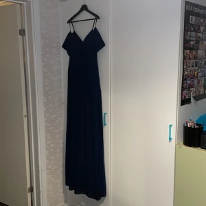 Marinblå långklänning med smala axelband - Säljer en marinblå långklänning/bal  med smala axelband och v-ringad omlottdesign. Klänningen har ett flytande fall, lätt volang vid axlarna och dold dragkedja i sidan. Perfekt för dig som vill ha en elegant look med enkel men snygg detalj.Jättevacker färg.
