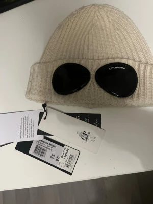 Vit mössa med goggles från C.P. Company - Säljer min vita mössa ifrån cp company, den är använd sparsamt och är i riktigt gott skick, mössan är one size!, köpt på NK för 1299kr, tags medföljer! 