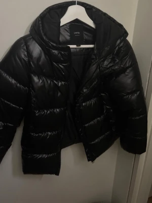Svart pufferjacka från LMTD XXS - Säljer en svart, glansig pufferjacka från LMTD i storlek XXS. Jackan har huva, quiltad design och dragkedja framtill. Perfekt för kalla dagar och riktigt snygg till alla outfits. Materialet är syntet och jackan har en loose passform.